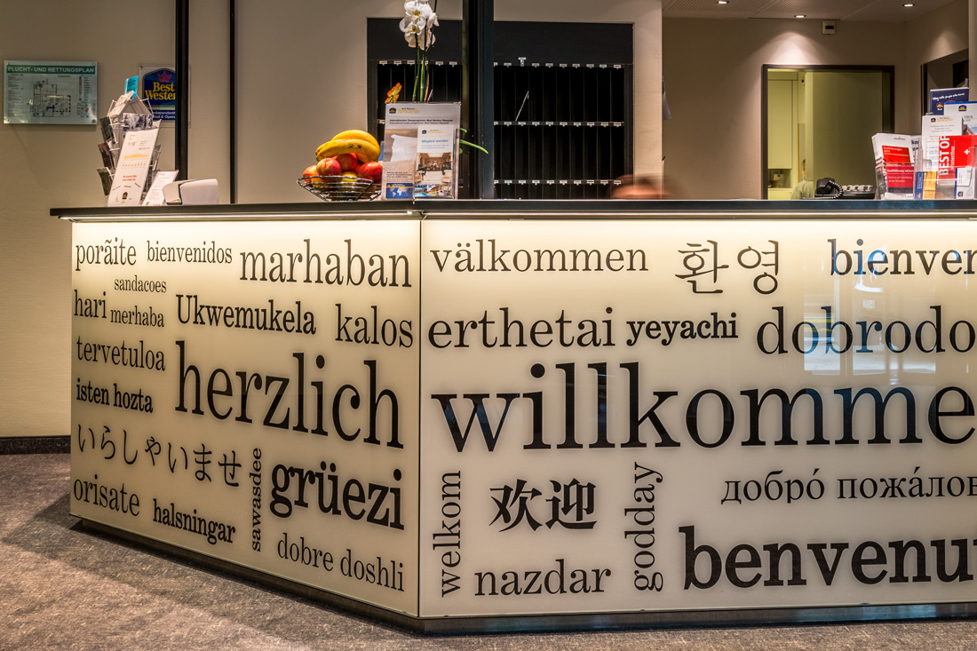 Beleuchtete Empfangstheke mit matten Glaswaenden, beschriftet mit schwarzen Schriftzügen: Willkommen in verschiedenen Sprachen