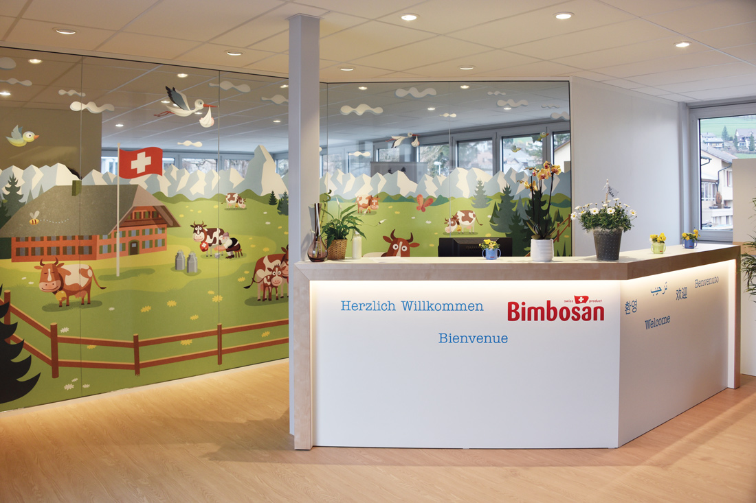 Showroom Empfang mit grossem Bild auf foliertem Glas: Schweizer Bauernhaus, Kuhweide und Bergpanorama in kindlich froehlichem Bauernmalereistil