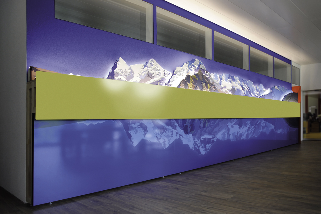 3D Wandgestaltung: Das Bergpanorama in 9 Metern Laenge, weisse Schneeberge, stylisierte gruene Wiese und Spiegelung im Bergsee in der unterenn Bildhaelfte