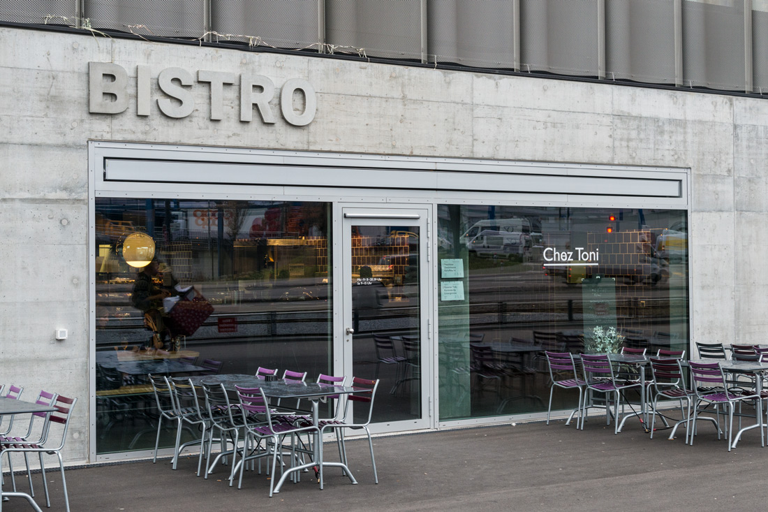 Aussenbeschriftung mit Betonbuchstaben schalungsglatte Oberflaeche, Zargen: Körnung Kies sichtbar. Aufnahme vor dem Bistro mit Tischen und Stuehlen.