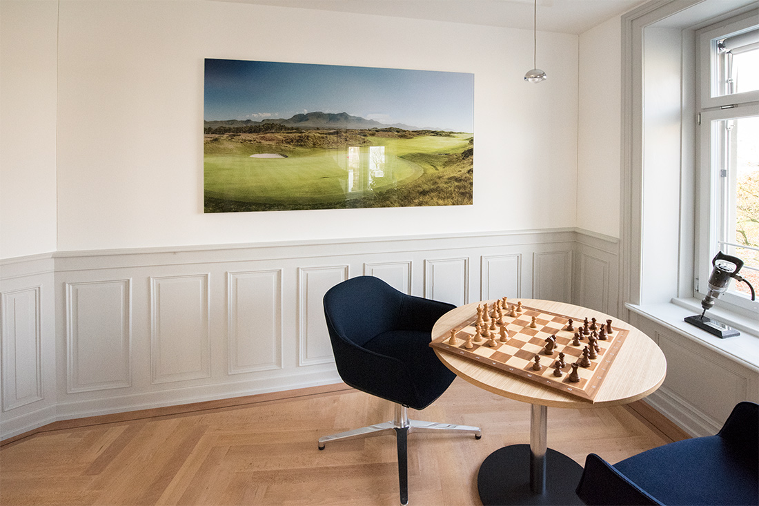Salontisch mit Schachbrett im hellen Raum, Glasbild mit Stimmungsbild Landschaft und Kassetten-Taefelung in hellem Lichtgrau an der Wand