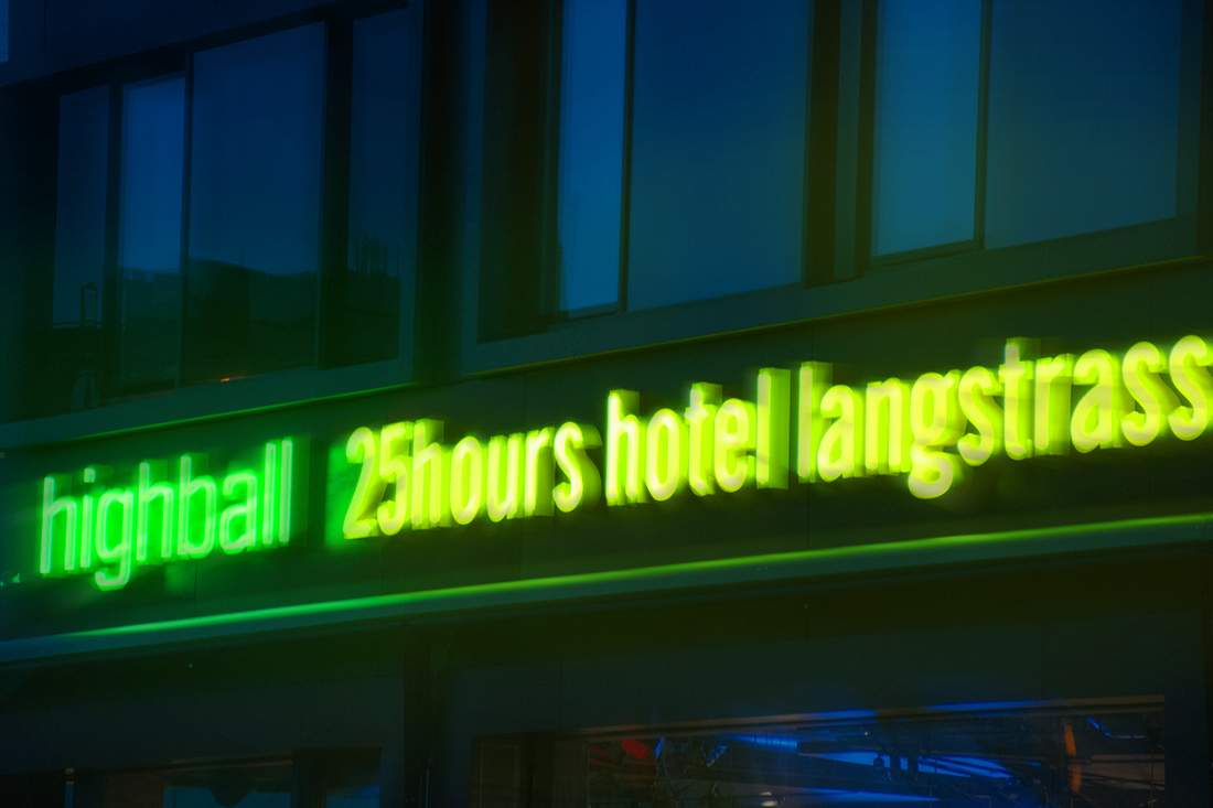 Nachtaufnahme: Neon-Schriftzuege an der Fassade des Hotels strahlen gelb und gruen im Dunkeln